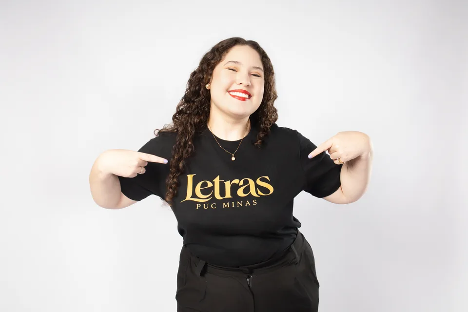 Bruna sorrindo e apontando para a camisa de Letras da PUC Minas no ensaio em estúdio