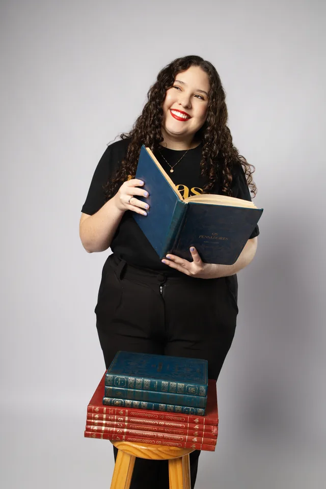 Bruna com um livro aberto e outros livros sobre a banqueta, usando a camisa de Letras.