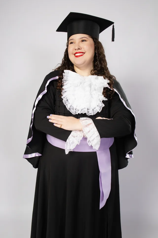 Bruna sorrindo de beca e capelo com faixa lilás no ensaio de formatura de Letras em BH
