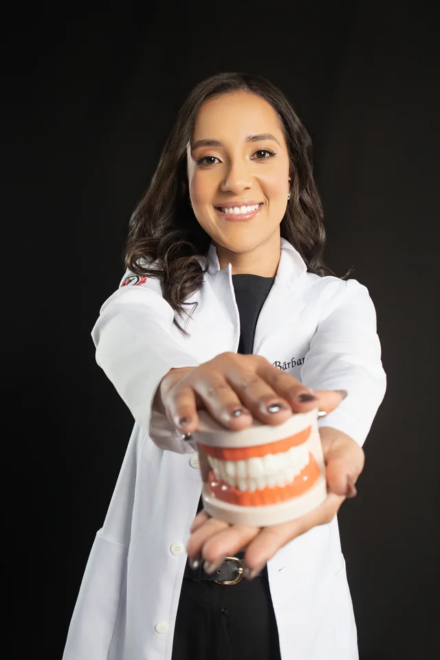 Bárbara com jaleco de Odontologia apresentando um modelo de arcada dentária sobre fundo escuro no estúdio