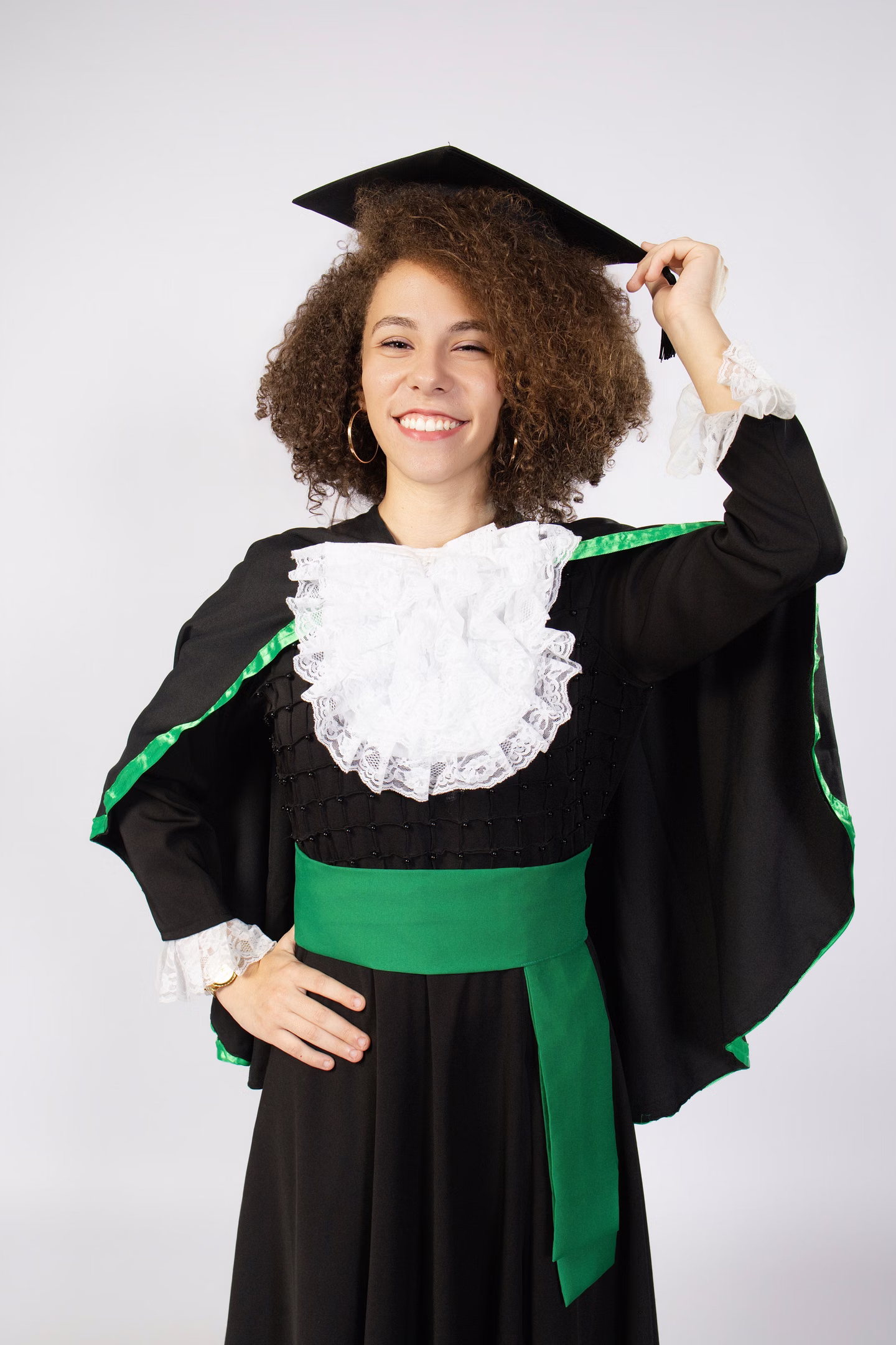 Laila com beca verde, sorrindo e tocando o capelo no ensaio de formatura em Belo Horizonte