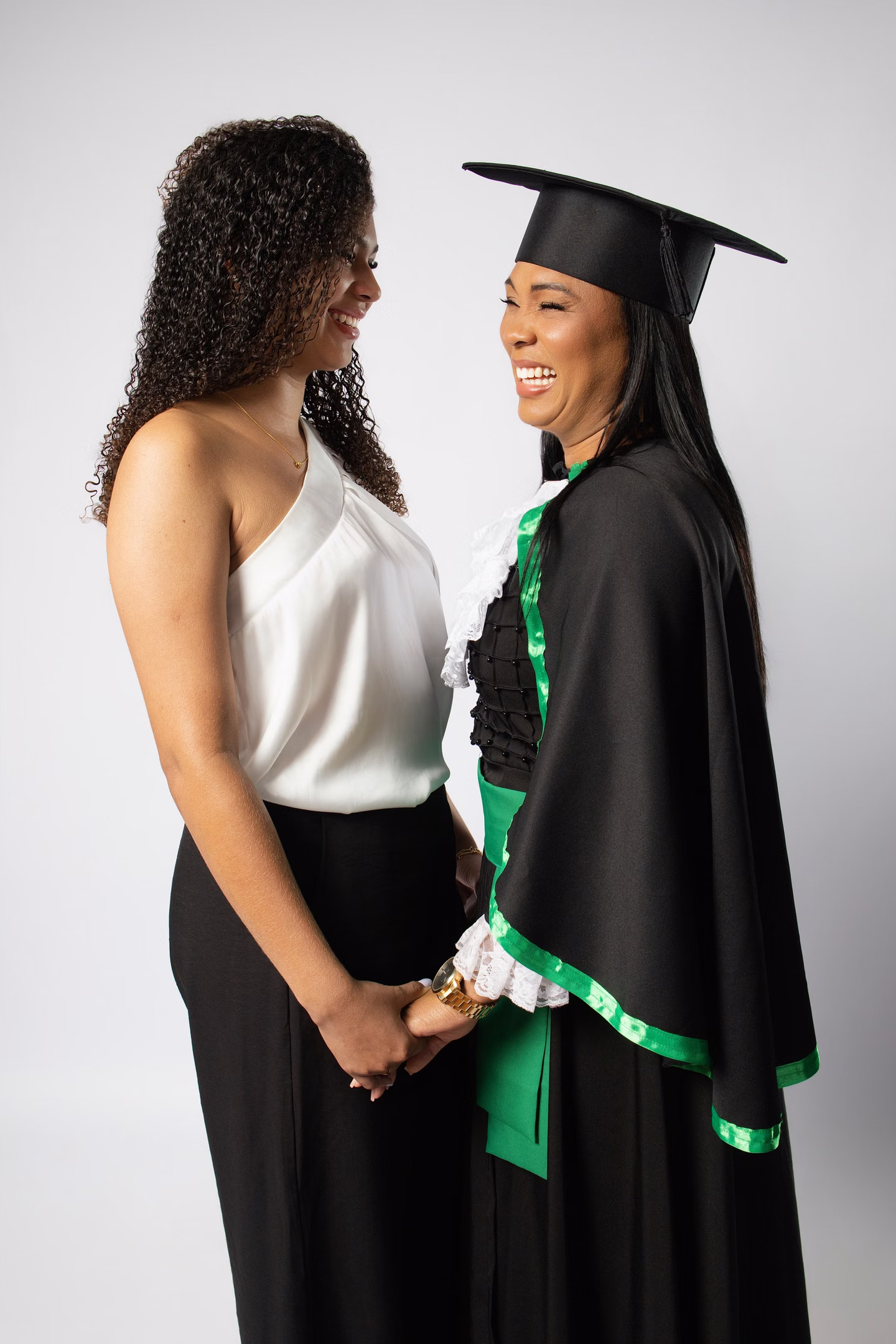 Lilian de beca e uma mulher de mãos dadas, sorrindo uma para a outra no ensaio de formatura em Belo Horizonte